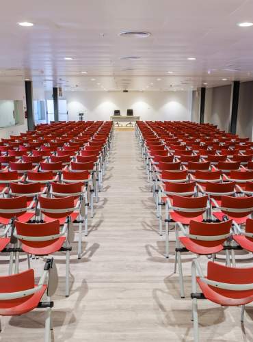 Uma sala de conferências preparada para um evento profissional em Ibiza, com filas de cadeiras vermelhas viradas para um palco com uma mesa para oradores, num espaço moderno equipado para conferências, seminários e conferências de imprensa.