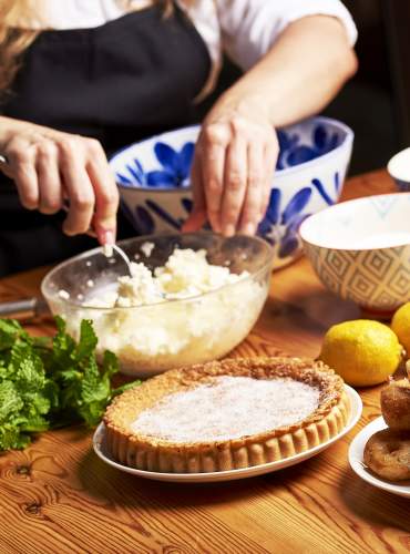 Uma pessoa a preparar uma sobremesa tradicional ibizenca numa mesa de madeira com ingredientes frescos como queijo, ovos, limões e hortelã, acompanhada por uma flaó (um tipo de salsicha) e algumas orelletas (bolinhos fritos) acabadas de preparar, representando a gastronomia tradicional de Ibiza.