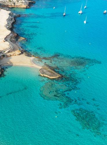 Vista aérea de Cala Comte, en Ibiza, con aguas turquesas y transparentes, pequeñas calas de arena blanca, un chiringuito junto al mar, embarcaciones fondeadas y un paisaje mediterráneo rodeado de pinares y costa rocosa.