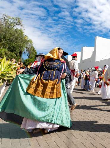 Um grupo de dançarinos com trajes tradicionais de Ibiza executa o *ball pagès* em frente a uma igreja branca decorada com bandeirolas durante um festival popular, rodeados por habitantes locais e visitantes sob um céu azul.