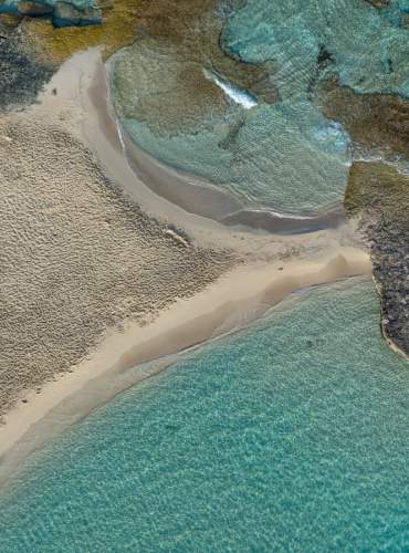 A praia de Ibiza vista do céu, com as águas turquesas e transparentes do Mar Mediterrâneo e uma grande rocha que se estende desde a areia até ao mar.