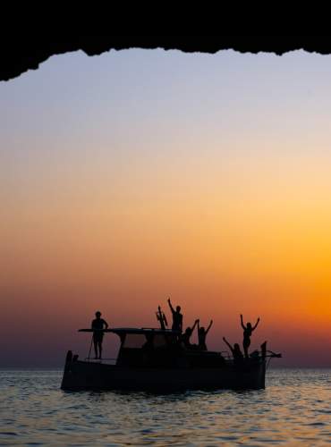 Um grupo de amigos acena de um barco contra a luz, num pôr-do-sol alaranjado em pleno mar em Ibiza.