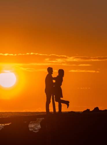 Um casal abraça-se numa praia de Ibiza durante um belo pôr-do-sol, com o céu alaranjado e as suas silhuetas escuras iluminadas por trás.