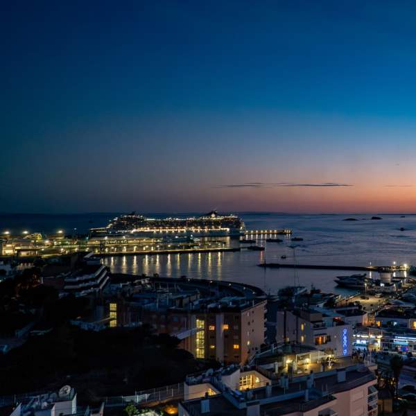 Vista panorâmica do porto de Ibiza ao entardecer, com um navio de cruzeiro atracado, uma marina repleta de barcos, a cidade iluminada e o contorno de Formentera no horizonte sob um céu em tons de azul e laranja.