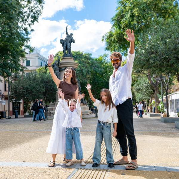 Imagen de una familia joven con dos niñas saludando al espectador. Están en el centro de la ciudad Ibiza, en el Paseo Vara de Rey. Al fondo se ve el monumento central de la plaza, en los laterales varios árboles y terrazas.