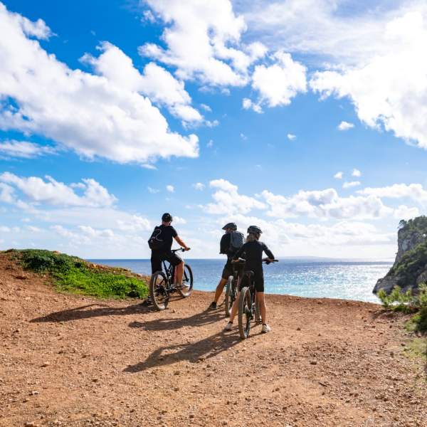 Tres personas recorren en bicicleta un sendero costero de Ibiza y se detienen en un mirador natural con vistas al mar Mediterráneo. El paisaje combina acantilados, vegetación mediterránea y aguas cristalinas bajo un cielo azul con nubes, mostrando una experiencia de cicloturismo en plena naturaleza.