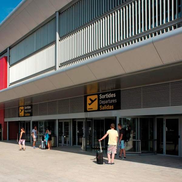 Fachada do Aeroporto de Ibiza com a zona de embarque, passageiros a entrar no edifício com bagagem e sinalização visível em espanhol, catalão e inglês; principal porta de entrada aérea da ilha.