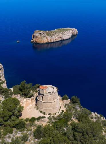 Vista aérea de la Torre des Molar, una histórica torre de defensa situada sobre un acantilado del norte de Ibiza, rodeada de pinar y con el islote de sa Conillera al fondo, sobre las aguas azul intenso del mar Mediterráneo.