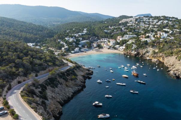 Vista aérea de Cala Vadella, en Ibiza, con una pequeña bahía de aguas turquesas y cristalinas donde fondean numerosas embarcaciones. La playa de arena se encuentra rodeada de colinas cubiertas de pinos y casas blancas escalonadas sobre la ladera, mientras una carretera serpentea junto a la costa rocosa.