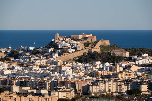 Vista panorámica de la ciudad de Ibiza con Dalt Vila elevándose sobre el casco urbano. Las murallas renacentistas, la catedral y las edificaciones históricas dominan una colina frente al mar Mediterráneo, mientras la ciudad moderna se extiende en primer plano bajo un cielo despejado.