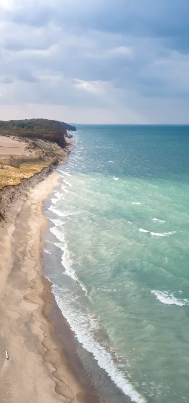 Aerial view of a lakeshore. The water on the right is a variety of light and dark blues. The dunes end in green vegetation and woods.