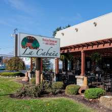 A stucco building with a fenced in patio sits near grass. The sky is blue. A sign out front reads La Cabaña and Mexican Restaurant and Cantina
