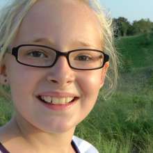 A blond girl with glasses is smiling. A vegetation-covered dune is in the background.