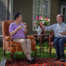 A man and women sit on furniture drinking tea while sitting on the front lawn.