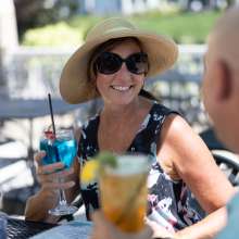 A woman in a hat and sunglasses holds a glass while smiling