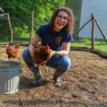 Woman kneeling down in a chicken coop, holding an orange chicken. A bucket sits next to her and another chicken is walking in the background.