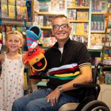 A young child stands next to a man sitting in a wheelchair. Both are smiling broadly and holding puppets.