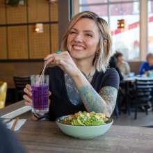 A woman with blonde hair and tattoos holds a purple drink while talking to someone unseen on the other side of the table.