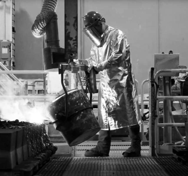 In a black and white industrial scene, a worker wearing a reflective protective suit and visor handles a large metal ladle, pouring molten material into a casting mold. The factory environment includes heavy equipment, chains, and vents, with smoke rising from the glowing liquid.