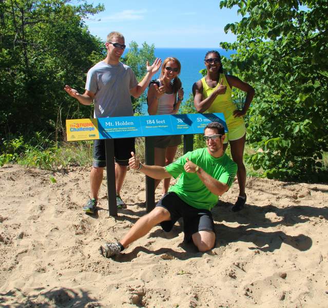 Four athletes stand atop a sand dune near a 3 Dune Challenge sign. The blue water of Lake Michigan is in the distance.