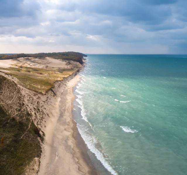 Aerial view of a lakeshore. The water on the right is a variety of light and dark blues. The dunes end in green vegetation and woods.