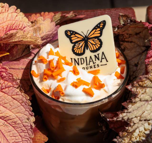A coffee drink with whipped cream, orange sprinkles, and a butterfly candy that says "Indiana Dunes" has plants around it.