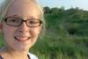 A blond girl with glasses is smiling. A vegetation-covered dune is in the background.
