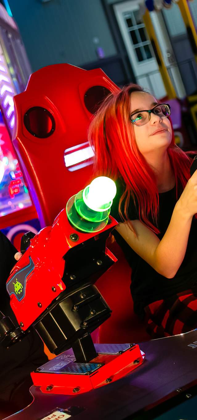 Two kids with reddish-pink hair sit in an arcade playing a shooting game