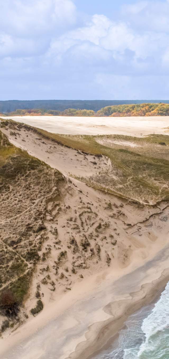 An aerial view of a massive dune with vegetation on it. The lake to the right is aqua with white wave crests.