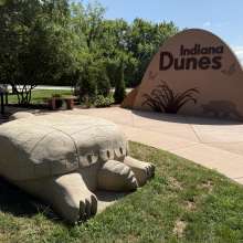 A large stone turtle sculpture at the Indiana Dunes Visitor Center, part of the Indigenous Cultural Trail, with a rock engraved with the Indiana Dunes logo in the background.