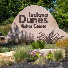 A dune-shaped sign says "Indiana Dunes Visitor Center" and has imagery of a sandhill crane and marram grass.