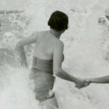 Vintage black-and-white photograph of a person wading through crashing waves while holding another person's hand.