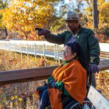 A brunette woman using a wheelchair looks at the fall colors in the distance. A park ranger points to something out of the picture.