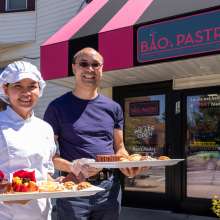 A chef and her husband stand in front of their pastry shop, holding trays of French pastries