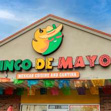 The exterior of a restaurant named "Cinco de Mayo," featuring a playful logo of a smiling character wearing a sombrero. The vibrant sign includes bold, colorful letters with Mexican-inspired patterns. Below the name, a sign reads "Mexican Cuisine and Cantina." The facade is adorned with brightly colored papel picado banners in various colors, adding a festive touch to the entrance. The building has a light-colored stucco finish with brick accents, and the sky above is a clear blue.