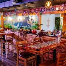 Bright and colorful interior of a restaurant decorated with vibrant murals depicting horses and landscapes on the walls. The space features wooden tables and chairs, with some tables adorned with intricate tile mosaics. Decorative lanterns hang from the ceiling, casting a warm glow. Small piñatas are suspended from above, adding a festive touch. A saddle is displayed on a wooden divider, enhancing the rustic and playful ambiance. Various potted plants and decor items are placed around, contributing to the lively atmosphere. A sign on one table indicates that patrons must be 21 or older to enter.