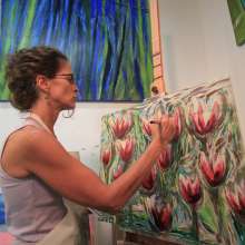 A woman in a tank top and apron paints flowers on a canvas. Other paintings hang on a white wall behind her.