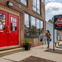 The exterior entrance of the Chesterton Art Center in Chesterton, Indiana, featuring striking red double doors set into a brick building with a gallery and classes sign. Two visitors walk along the sidewalk past potted flowers at the welcoming entryway.