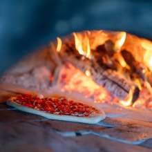 A pizza sits in a brick oven in front of logs that are aflame.