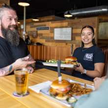 A waitress holds a plate with a burger on it in front of a table with two customers.