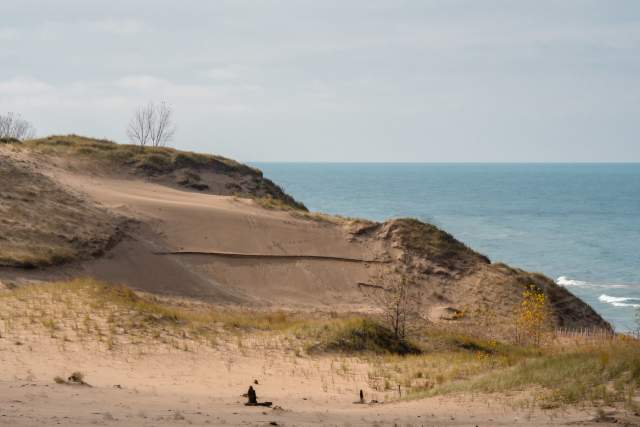 Sandy dunes with sparse vegetation overlooking a large body of water under a cloudy sky.