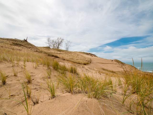 Sandy dunes with sparse green and brown grass under a partly cloudy sky, with a glimpse of the ocean on the right.