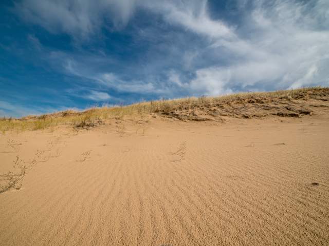 A sandy dune with sparse vegetation under a blue sky with wispy white clouds.