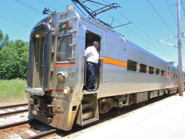 A conductor stands in the doorway atop steps on a silver train.