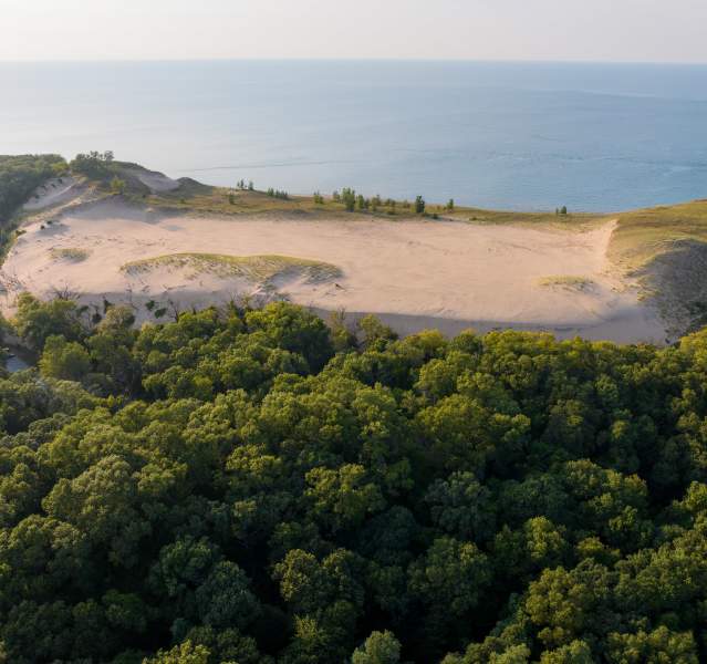Aerial view of a large sand dune surrounded by green trees, with a calm blue lake in the background.