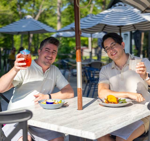 Two people toasting drinks at an outdoor patio table at The Goblin and the Grocer restaurant in Beverly Shores, Indiana