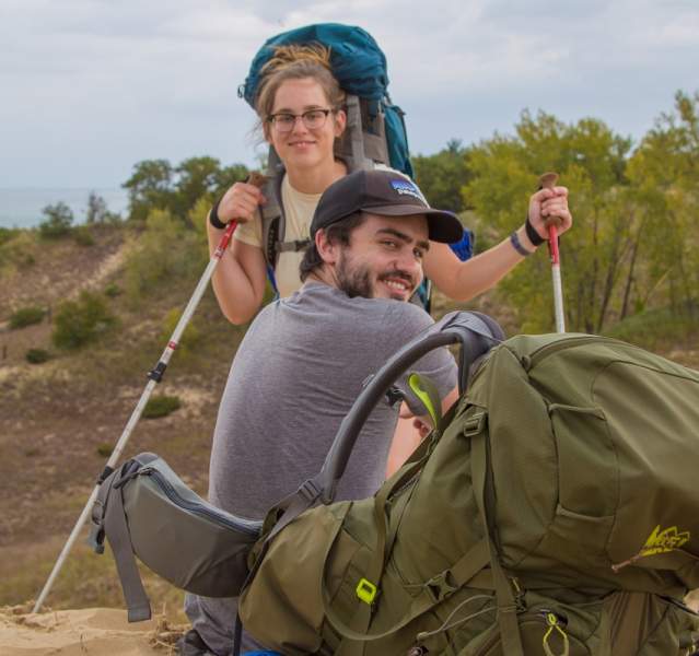 Two hikers pause on sandy terrain, equipped with trekking poles and backpacks, overlooking a serene lake and lush greenery.