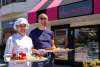 A chef and her husband stand in front of their pastry shop, holding trays of French pastries