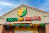 The exterior of a restaurant named "Cinco de Mayo," featuring a playful logo of a smiling character wearing a sombrero. The vibrant sign includes bold, colorful letters with Mexican-inspired patterns. Below the name, a sign reads "Mexican Cuisine and Cantina." The facade is adorned with brightly colored papel picado banners in various colors, adding a festive touch to the entrance. The building has a light-colored stucco finish with brick accents, and the sky above is a clear blue.