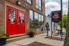 The exterior entrance of the Chesterton Art Center in Chesterton, Indiana, featuring striking red double doors set into a brick building with a gallery and classes sign. Two visitors walk along the sidewalk past potted flowers at the welcoming entryway.